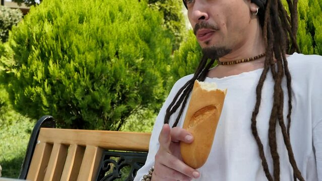 A young adult guy with dreadlocks and an earring in his ear sitting on a park bench uses a laptop for a video call and eats a fresh loaf of bread biting and talking