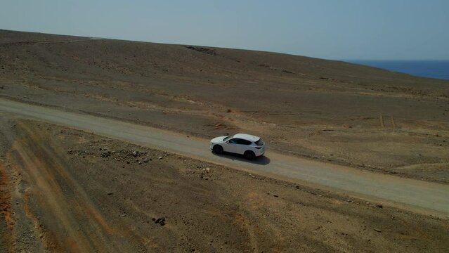 Aerial view laterally following a white car on a volcanic and desert road on the island of Fuerteventura on a sunny day.