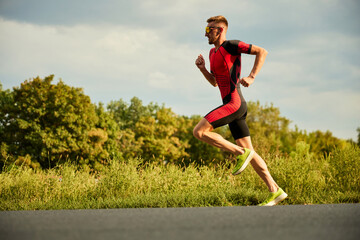 Fototapeta premium Dynamic image of competitive man in sportswear, athlete training outdoors on summer day, running. Marathon. Concept of professional sport, triathlon preparation, competition, athleticism