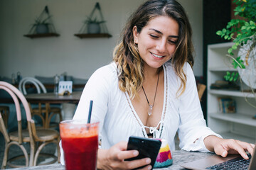 Cheerful woman watching mobile phone and typing on laptop in cafe