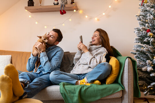 Loved Ones. Happy Young Couple And Their Small Dog Together At Home During The Christmas Holidays. Woman Takes A Photo Of Her Boyfriend Kissing Their Dog.