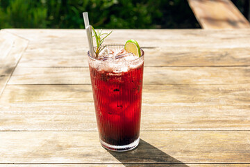 Fresh homemade mulberry ice tea with rosemary and lemon served on wooden table outdoors. summer cold fruit drink in sunny afternoon with green garden behind