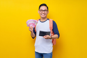 Smiling young Asian student man in casual clothes and glasses backpack holding cash money and...