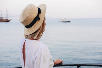 Woman in white dress and straw hat looking at the sea with boats 
