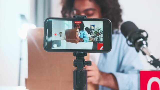 Young energetic curly Arabian man recording blog with review of new gadget for high-quality video shooting and working as operator sits in room near smartphone mounted on tripod. Unboxing vlog