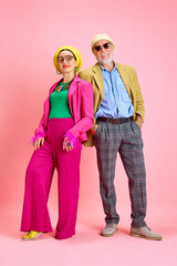 Full-length portrait of beautiful senior couple, elegant man and woman in stylish clothes standing against pink studio background. Concept of beauty and fashion, relationship, modern style, age