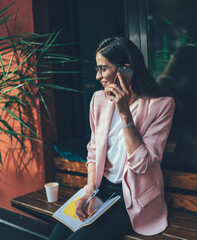 Joyful adult woman with takeaway hot drink and notebook talking on phone in city