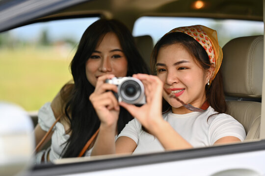 Two happy female friends taking photos from a car window with vintage retro cameras, enjoying weekend road trip together