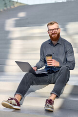 Handsome red-haired man with a beard sits on the stairs during a break with a laptop