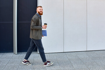 A man with a red beard dressed in a shirt and jacket drinks coffee while walking