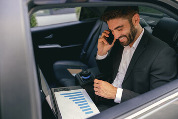 Handsome businessman in suit using personal computer and making phone call in taxi