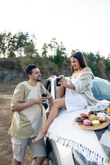 Couple enjoying picnic time on the sunset in a meadow, standing near suv car.