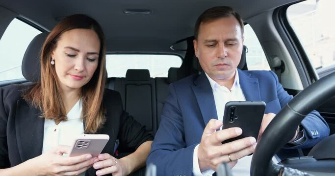 Office workers man and woman in suits sit in car holding phones. Colleagues check social media news talking. Businesswoman smiles reading messages on phone