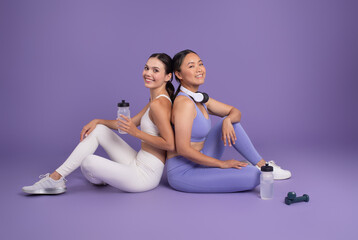 Fototapeta na wymiar Diverse ladies friends resting with water bottles after workout, sitting on purple studio background