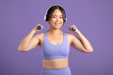 Woman happily exercising with hand dumbbells and listening music in headphones, purple studio