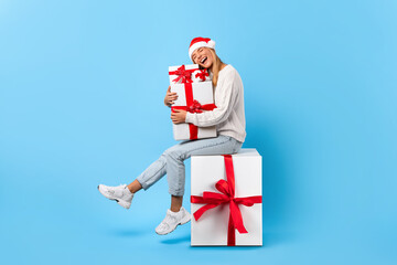 Excited woman posing with many Christmas presents and sitting on big gift box, lady wearing Santa hat, blue background