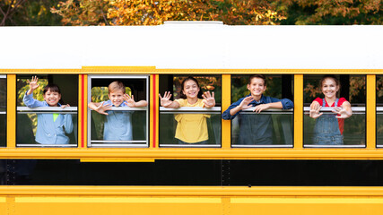 Joyful kids looking out from yellow school bus, waving hands at camera