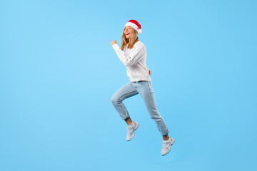 Christmas rush. Happy lady in Santa hat jumping on blue background, excited woman running to buy...