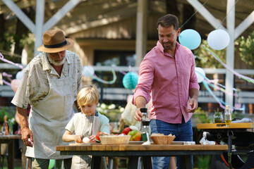 Young boy cutting vegetables for grilling. Father, grandfather and son grilling together at garden BBQ. Three generations of men at summer family garden party.