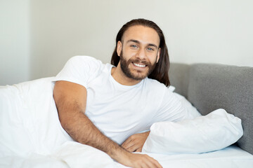 Smiling Pleased Long-Haired Man Lying In Comfortable Bed