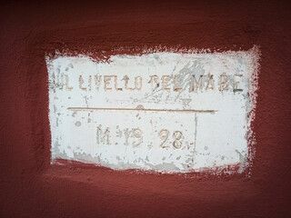 Scritta incisa sul muro di un edificio che indica la posizione SUL LIVELLO DEL MARE metri 19,28, sfondo bianco e bordeaux 