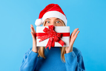 Young lady in Santa hat hiding face with New Year present over blue studio background