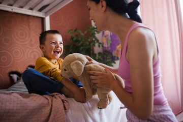 Single mother waking her son up for school in the morning. Mom awakens the schoolboy with a stuffed toy, playing and making him laugh.