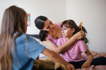 Mother with two daughters on a bed in the bedroom. Mother combs daughter's long hair with comb.Concept of Mother's Day and maternal love.