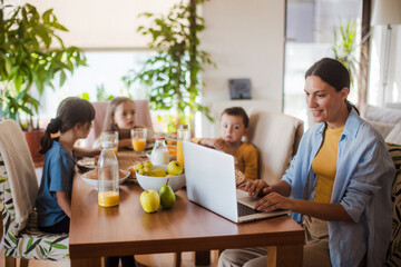 Mother working on laptop at kitchen table while children have breakfast. Remote work and home office for mothers with children.