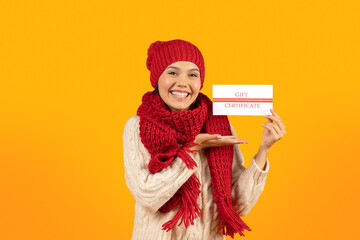 Lady in winter knitwear showing shopping gift certificate, yellow background