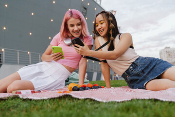 Two cheerful girls using smartphones while sitting on grass outdoors