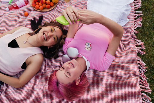 Top view of two cheerful girls taking selfies while laying on blanket in park