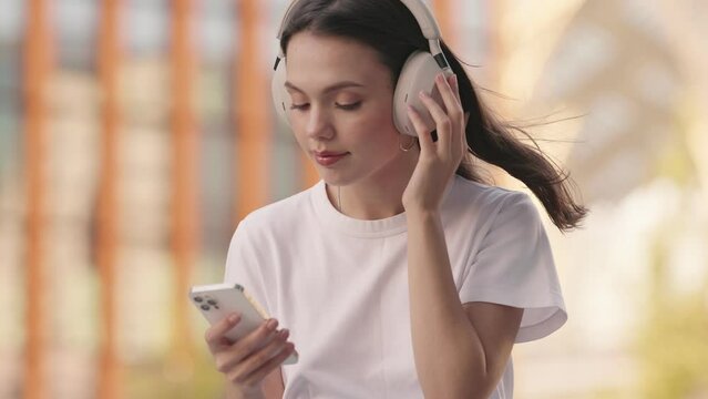 A young woman happily listens to music with noise-isolating headphones, scrolling through her favorite playlist on her smartphone while using touch gestures on her headphones