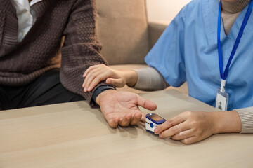 Close-up nurse visits an elderly person for a health check. A young nurse examine pulse and oxygen saturation at home. Happy elderly Asian man in his 60s measuring blood pressure in his home.