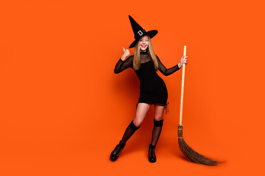 Full size photo of pretty blonde teenager girl hold broom thumb up dressed black halloween outfit isolated on orange color background