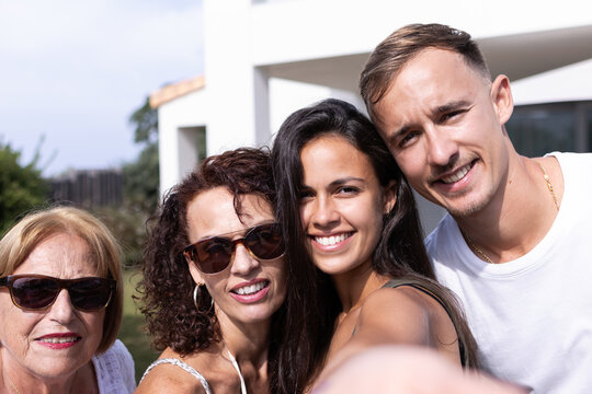 A smiley photo of a caucasian family taking a selfie of themselves, looking at camera and with their home behind them