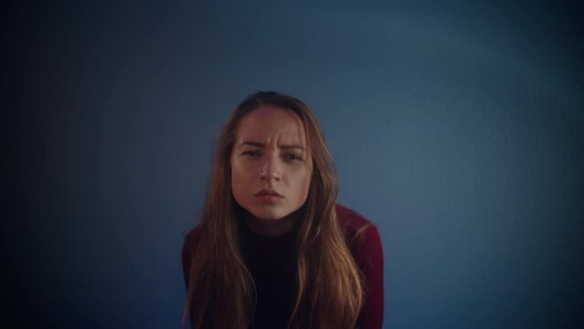 Portrait of a young caucasian girl with long brown hair in a burgundy sweater looking at the camera and trying to see something in front of her on a blue background. Emotion of learning and interest