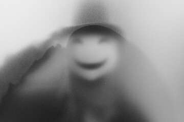 Bangkok, Thailand - August, 18, 2023 : Mysterious man in smiling mask stands behind frosted glass, spooky style, blurred image. Halloween concept. - 662727534