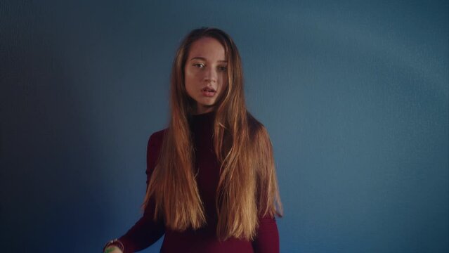 Portrait of a young beautiful erotic attractive caucasian girl in a burgundy sweater looking at the camera on a blue background. She seductively unbinding and playing with her long hair in slow motion