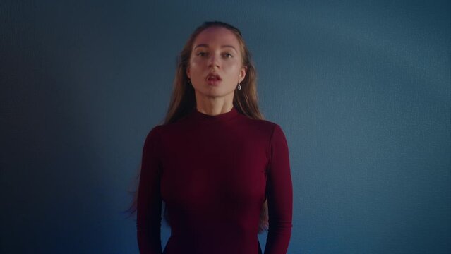 Portrait of a young beautiful erotic attractive caucasian girl in a burgundy sweater looking at the camera on a blue background. She lifts her head and her long brown hair falls down in a slow motion
