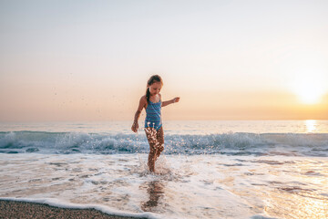 Fototapeta na wymiar The child happily plays in the waves on the seashore. A girl in a blue swimsuit laughs while playing in the waves on the beach