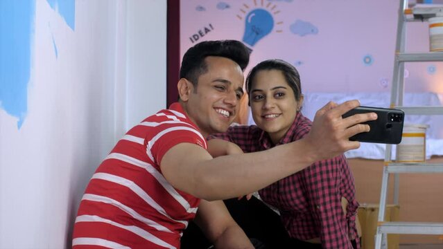A cheerful Indian couple clicking a selfie together in their new rented apartment - moving to new house. A young man and woman posing while taking pictures on mobile while sitting on the floor agai...