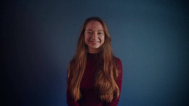 Portrait of a young beautiful smiling pretty caucasian girl with long brown hair in a burgundy sweater looking at the camera on a blue background. Emotion of joy and satisfaction