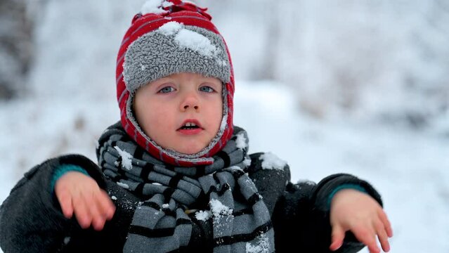 little boy in a hat with earflaps and a fur coat rides on a retro sleigh in a magical winter forest and sleds. Fairytale Christmas greeting card with baby. Baby in winter snowy forest eats snowflakes