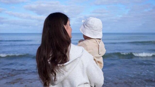 Brunette mama carefully holds sweet daughter in arms watching sea waves. Woman kisses little girl putting tenderness with maternal in kiss