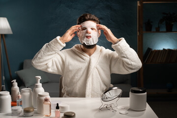 Bearded young man in bathrobe taking off moisturizing sheet mask. Face care and beauty treatments for man.