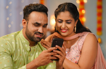 Happy young Indian couple using mobile phone together at home during diwali festival celebration in front of gifts- concept of social media sharing, festive wishes or greetings and Joyful Connection