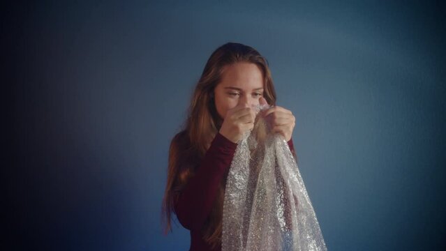 Portrait of young beautiful playful happy caucasian girl with long brown hair in burgundy sweater looking at the package and breaking bubble wrap on blue background. Concept of calm and anti-stress