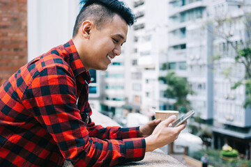 Joyful youthful Asian male having fun with cellphone on balcony
