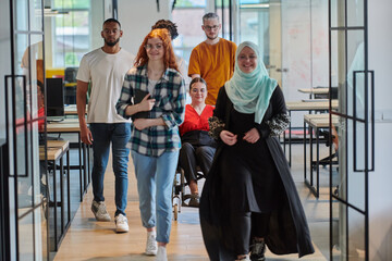 A diverse group of business people walking a corridor in the glass-enclosed office of a modern startup, including a person in a wheelchair and a woman wearing a hijab, showing a dynamic mix of